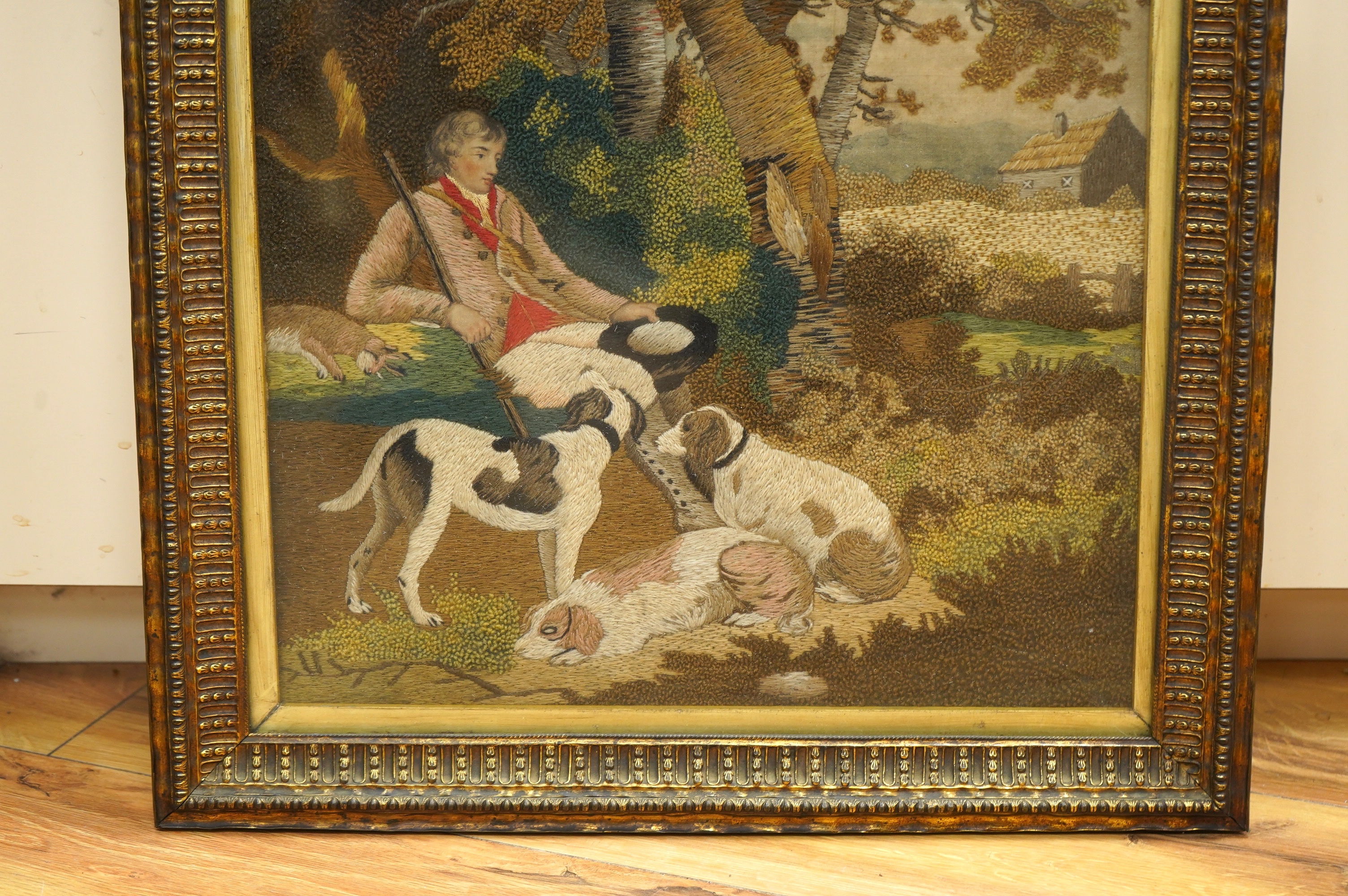 A framed early 19th century wool worked embroidery of huntsman sitting with a rabbit and his three hunting dogs under a tree, by a stream, with a field and cottage in the background, the embroidery worked in fine multi-c